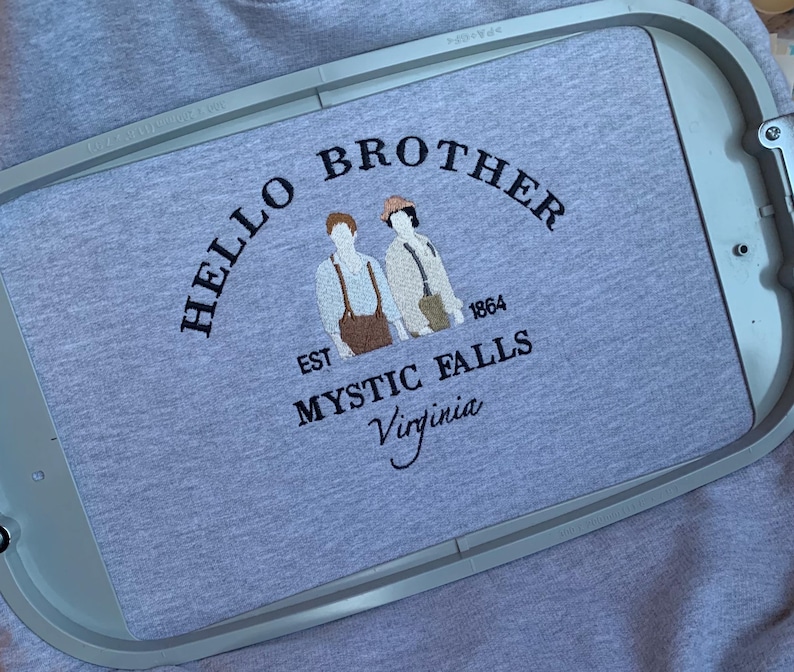 May include: A light gray t-shirt with the embroidered text "HELLO BROTHER" above two figures and the words "EST 1864 MYSTIC FALLS Virginia". The design is in dark brown thread.