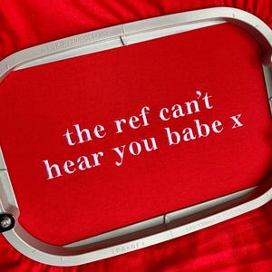 May include: A bright red fabric held in a light grey embroidery hoop measuring 180x130mm. The fabric features white embroidered text that reads: the ref can't hear you babe x. The lettering is centred and clearly visible against the red background.