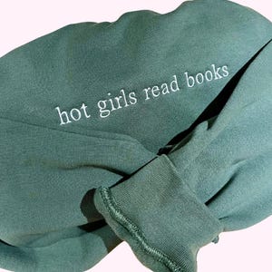 Può includere: Una felpa verde foresta con la scritta ricamata in bianco "hot girls read books". La felpa è piegata, con il polsino della manica visibile. Lo sfondo è rosa chiaro.