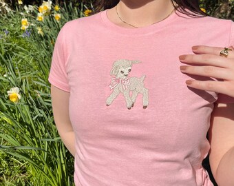 Peut inclure: Un t-shirt rose avec un agneau blanc brodé portant un nœud rose. L'agneau est tourné vers la gauche de l'image.