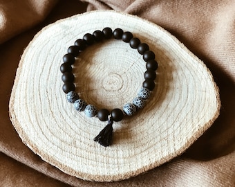 May include: A black and white beaded bracelet with a black tassel. The bracelet is made of round beads and is on a wooden surface.