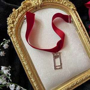 May include: A red velvet choker necklace with a silver guillotine pendant. The necklace is displayed on a white surface within a gold ornate frame. The background is black with white flowers and red rose petals.