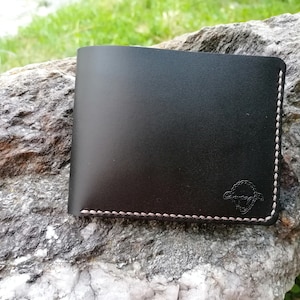 Puede incluir: Una cartera de cuero negro con un borde cosido blanco y un pequeño logotipo de oveja estampado en la parte delantera.