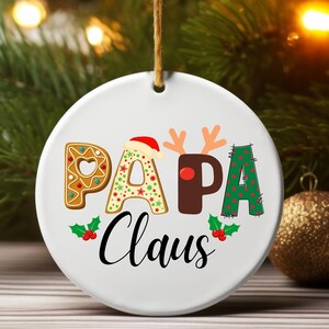 May include: White ceramic Christmas ornament with the text "Papa Claus" in black. The letters are decorated with gingerbread, reindeer antlers, and a red and green polka dot pattern.