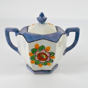 May include: A white porcelain sugar bowl with a blue lid and blue handles. The bowl is decorated with a floral design in orange, pink, and green.