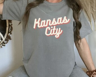 May include: A gray t-shirt with the text "Kansas City" in a retro-style font with a red outline. The text is in a cream color.