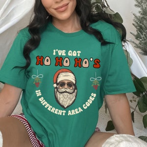 May include: Green t-shirt with a Christmas-themed graphic. The graphic features a cartoon Santa Claus with sunglasses and the text "I've got Ho Ho Ho's in different area codes".