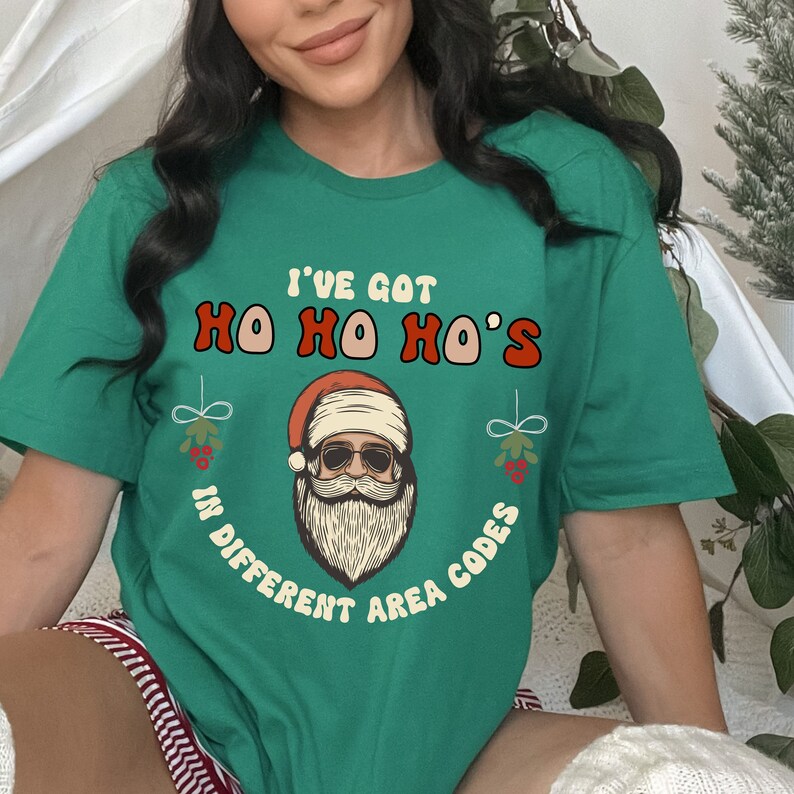 May include: Green t-shirt with a Christmas-themed graphic. The graphic features a cartoon Santa Claus with sunglasses and the text "I've got Ho Ho Ho's in different area codes".