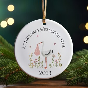 May include: White ceramic Christmas ornament with a stork carrying a pink bundle, surrounded by green and pink flowers. The text "A Christmas Wish Come True 2023" is printed on the ornament.