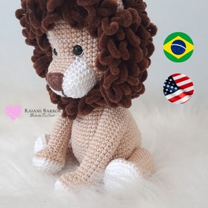 Pode incluir: Um leão de pelúcia de crochê em tons de bege, marrom e branco. O leão tem uma juba marrom fofa, um corpo bege e detalhes brancos nas patas e no focinho. O brinquedo está sentado em uma superfície branca e fofa.