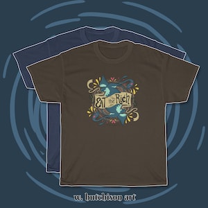 May include: Two t-shirts, one brown and one navy blue, with a graphic design of two blue birds in a nest with the text "EAT the Rich" on a banner. The graphic is surrounded by yellow, orange, and pink floral elements. The text "w. butchison art" is at the bottom of the image.