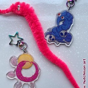 May include: A vibrant pink fuzzy worm toy with googly eyes, and three acrylic keyrings. The keyrings feature a blue bird with skulls, a pink crescent moon with wings, and a star. The artist's name, w. hutchison art, is in the bottom right corner.