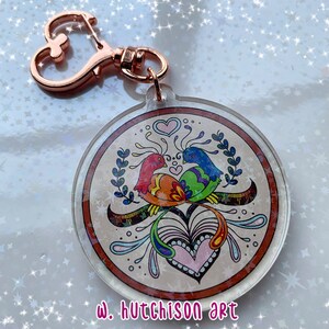 May include: A round, clear acrylic keychain with a colorful illustration of two birds intertwined with a heart and floral designs. The keychain features a rose gold metal heart-shaped clasp. The text "w. hutchison art" is printed at the bottom of the keychain.
