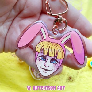 以下が含まれることがあります： ピンクと白のウサギの耳のキーホルダー。青い目の漫画の顔とピンクと白のウサギの衣装をしています。キーホルダーは透明なアクリル製で、金色の金属製のリングが付いています。