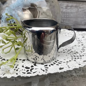 May include: A shiny silver creamer with a rounded body and a handle. The creamer is sitting on a white lace doily.