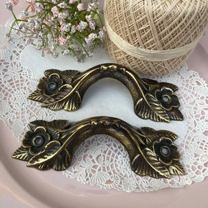 May include: Two antique brass drawer pulls with floral and leaf detailing. The pulls are arched and have a dark patina. The image also includes a ball of beige twine, a sprig of pink and white flowers, and a white lace doily.