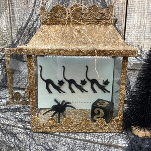 Puede incluir: Una linterna dorada de Halloween con una parte superior decorativa, que presenta una ara&ntilde;a negra y una tarjeta con siluetas de gatos negros. Tambi&eacute;n se ve un dado negro con el n&uacute;mero 2.