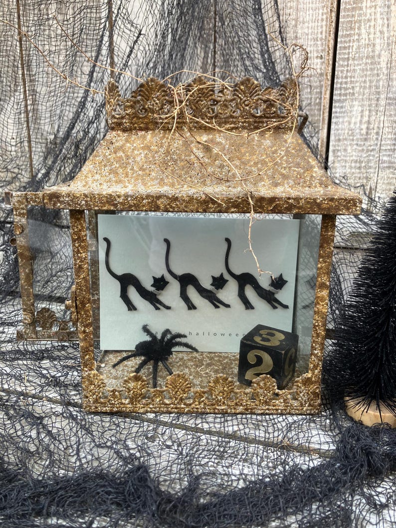 Puede incluir: Una linterna dorada de Halloween con una parte superior decorativa, que presenta una ara&ntilde;a negra y una tarjeta con siluetas de gatos negros. Tambi&eacute;n se ve un dado negro con el n&uacute;mero 2.