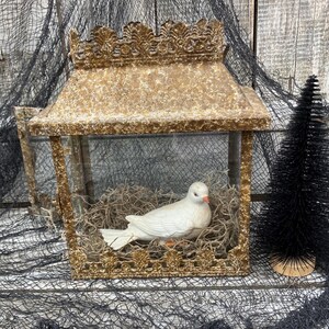 Puede incluir: Una linterna decorativa dorada con una paloma de cer&aacute;mica blanca en su interior, posada sobre un lecho de musgo seco. La linterna tiene un dise&ntilde;o detallado y ornamentado y un peque&ntilde;o &aacute;rbol negro a la derecha.