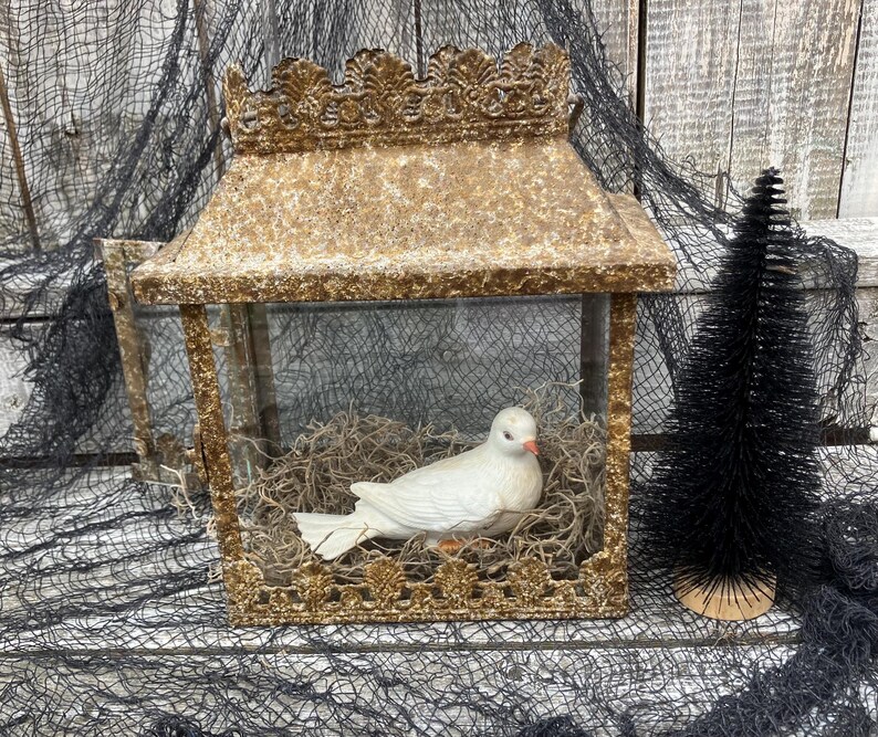 Puede incluir: Una linterna decorativa dorada con una paloma de cer&aacute;mica blanca en su interior, posada sobre un lecho de musgo seco. La linterna tiene un dise&ntilde;o detallado y ornamentado y un peque&ntilde;o &aacute;rbol negro a la derecha.