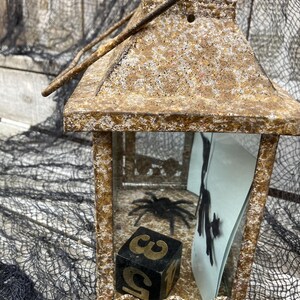 Puede incluir: Una linterna decorativa de metal envejecido con asa. En el interior, se ve una ara&ntilde;a negra y un dado con los n&uacute;meros 3 y 5. La linterna tiene un aspecto texturizado y envejecido, perfecta para Halloween.