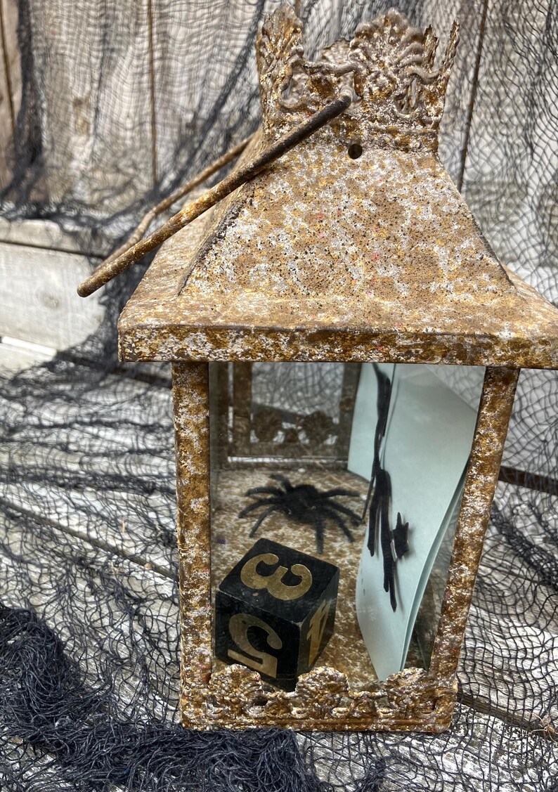 Puede incluir: Una linterna decorativa de metal envejecido con asa. En el interior, se ve una ara&ntilde;a negra y un dado con los n&uacute;meros 3 y 5. La linterna tiene un aspecto texturizado y envejecido, perfecta para Halloween.
