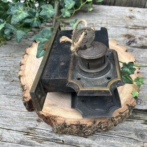 May include: An antique door lock set with a key attached by twine. The lock is made of dark metal with brass accents. The key is in the lock. The set is displayed on a wooden surface.