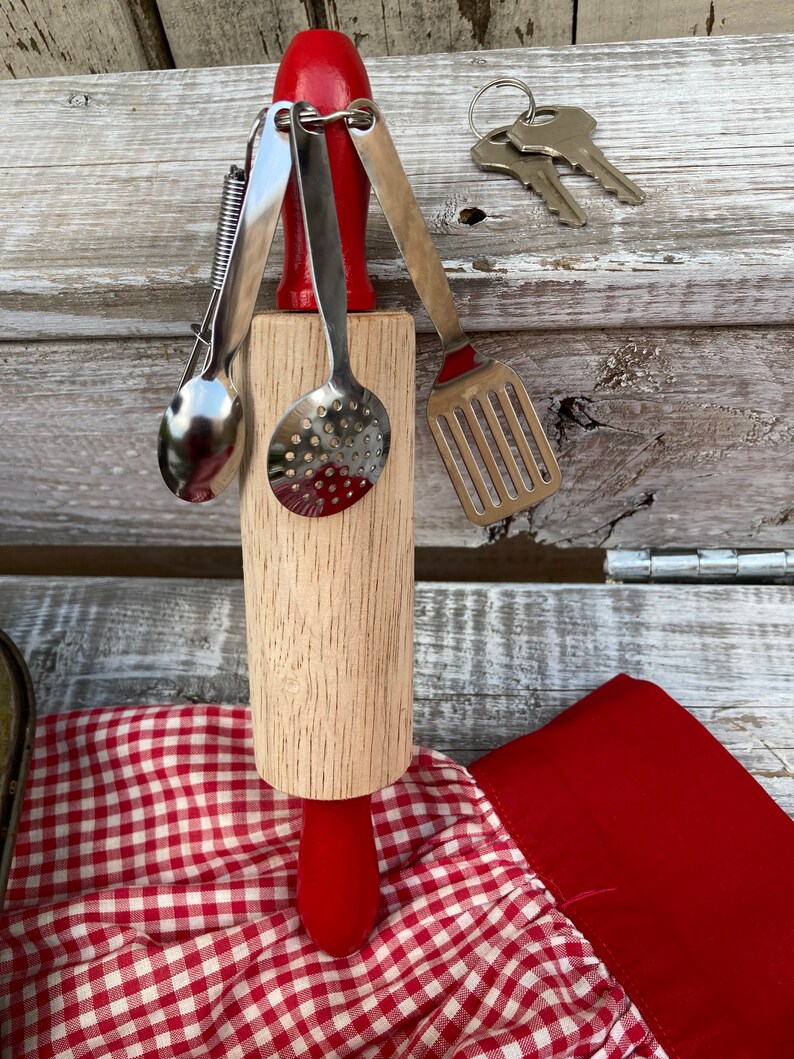 Op de afbeelding: Een houten deegroller met een rode handgreep en een set miniatuur keukengerei bevestigd aan een sleutelhanger. Het keukengerei omvat een lepel, een geperforeerde lepel, een spatel en een tang.
