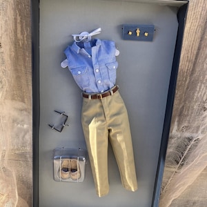 May include: A blue and white button-down shirt with a pair of khaki pants, a pair of brown shoes, and a small jewelry box with gold accents. The items are displayed in a black box with a white background. To remove clothing from box, lift package insert and carefully clip threads located on the reverse side.