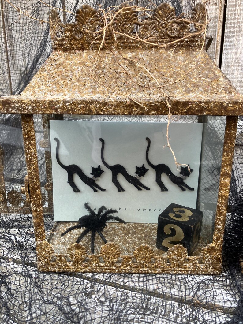 Puede incluir: Una linterna decorativa de estilo antiguo con acabado dorado, con tem&aacute;tica de Halloween. En el interior, una tarjeta muestra siluetas de gatos negros, una ara&ntilde;a peluda y un dado negro con el n&uacute;mero 2. Tambi&eacute;n es visible la palabra "halloween".