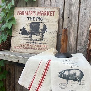 May include: A rustic wooden sign with the text "FARMER'S MARKET THE PIG" and a black and white illustration of a pig. Two cream-colored kitchen towels with red stripes and a pig illustration are also visible.