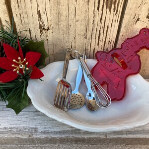 Op de afbeelding: Een witte keramische schaal met een rode plastic gingerbread koekjesvormer, een zilveren garde, een zilveren lepel en een zilveren geperforeerde lepel.