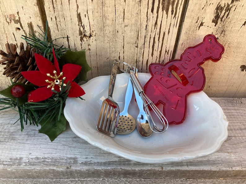 Op de afbeelding: Een witte keramische schaal met een rode plastic gingerbread koekjesvormer, een zilveren garde, een zilveren lepel en een zilveren geperforeerde lepel.