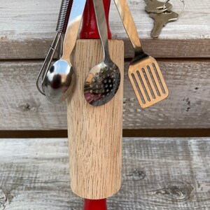 Op de afbeelding: Een houten deegroller met een rode handgreep en een set miniatuur keukengerei bevestigd met een metalen ring. Het keukengerei omvat een garde, een lepel, een geperforeerde lepel en een spatel.