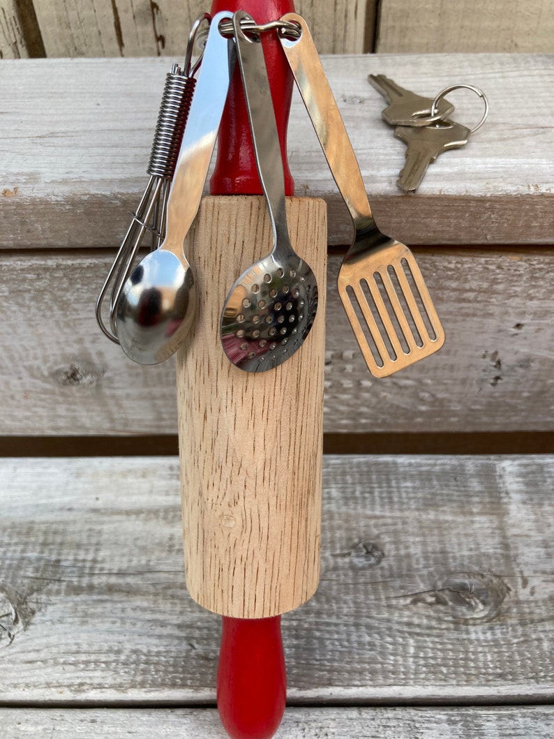 Op de afbeelding: Een houten deegroller met een rode handgreep en een set miniatuur keukengerei bevestigd met een metalen ring. Het keukengerei omvat een garde, een lepel, een geperforeerde lepel en een spatel.