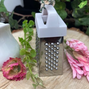 May include: A small, silver grater with a white plastic handle and a green and white fabric loop. The grater is sitting on a wooden surface.