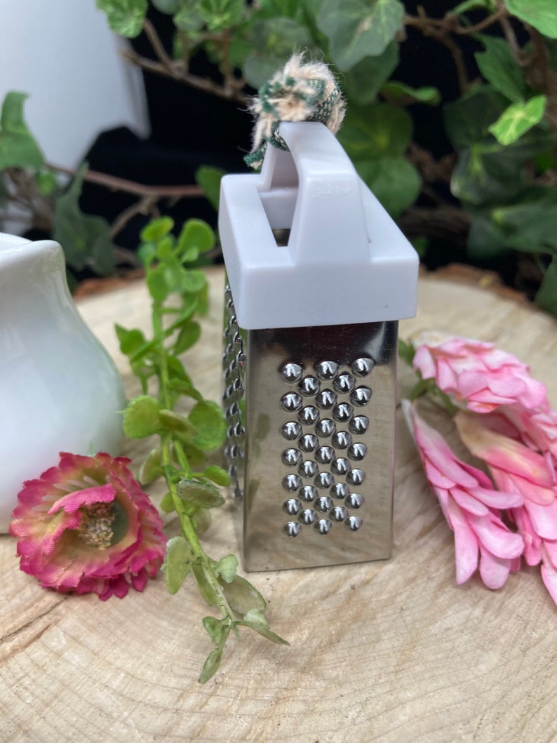May include: A small, silver grater with a white plastic handle and a green and white fabric loop. The grater is sitting on a wooden surface.