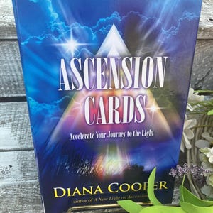 May include: A book titled "ASCENSION CARDS" with a blue cover featuring a white pyramid and bright light. The text "Accelerate Your Journey to the Light" is below the title. The author's name, Diana Cooper, is at the bottom.