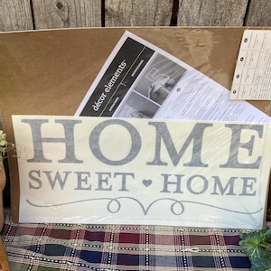 May include: A rectangular package containing a grey vinyl wall decal with the text HOME SWEET HOME and a small heart symbol. The decal is accompanied by a white instruction sheet labeled décor elements and a product information sticker on the back.