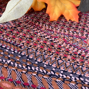 May include: Close-up of a patterned fabric with a mix of colors. The design features a combination of brown, red, and white geometric shapes and lines on a dark background. Autumn leaves are in the background.