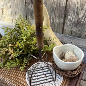May include: An antique potato masher with a wooden handle and metal head, resting on a white lace doily. A small white bowl with an egg and a nest of twigs sits nearby, with green foliage in the background.