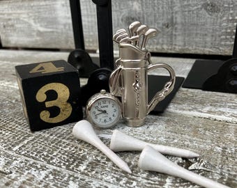 May include: A collection of golf-themed items, including a miniature silver golf bag with clubs, a small clock, white golf tees, and a black wooden block with the number 3 in gold. The items are arranged on a weathered wooden surface.