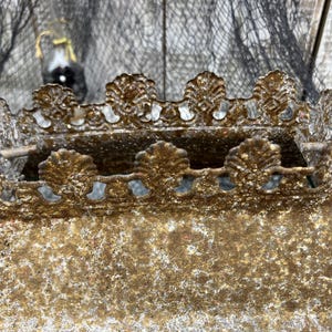 Puede incluir: Primer plano de una corona decorativa de metal de estilo antiguo. La corona es de color dorado deslucido con detalles ornamentados, incluyendo bordes festoneados y detalles en forma de hoja. El metal tiene un aspecto texturizado y envejecido.