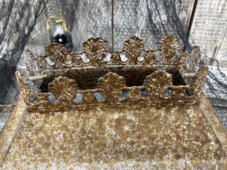 Puede incluir: Primer plano de una corona decorativa de metal de estilo antiguo. La corona es de color dorado deslucido con detalles ornamentados, incluyendo bordes festoneados y detalles en forma de hoja. El metal tiene un aspecto texturizado y envejecido.