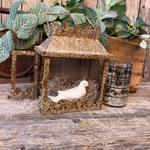 Puede incluir: Una linterna dorada de estilo vintage con una figura de paloma blanca en su interior, posada sobre un lecho de paja. La linterna est&aacute; decorada con una parte superior en forma de corona. Un vaso decorativo con un estampado floral negro est&aacute; junto a la linterna.