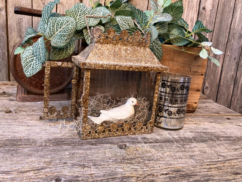 Puede incluir: Una linterna dorada de estilo vintage con una figura de paloma blanca en su interior, posada sobre un lecho de paja. La linterna est&aacute; decorada con una parte superior en forma de corona. Un vaso decorativo con un estampado floral negro est&aacute; junto a la linterna.