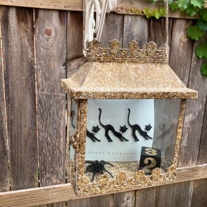 Puede incluir: Una linterna decorativa de estilo antiguo con un acabado dorado y bronce. La linterna presenta una parte superior y una base decorativas, con paneles de vidrio. En el interior, se ve una tarjeta con siluetas de gatos negros y el texto "happy halloween", junto con una ara&ntilde;a negra y un bloque con el n&uacute;mero 32.