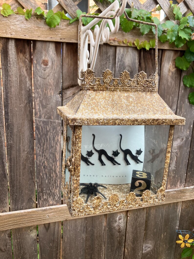 Puede incluir: Una linterna decorativa de estilo antiguo con un acabado dorado y bronce. La linterna presenta una parte superior y una base decorativas, con paneles de vidrio. En el interior, se ve una tarjeta con siluetas de gatos negros y el texto "happy halloween", junto con una ara&ntilde;a negra y un bloque con el n&uacute;mero 32.