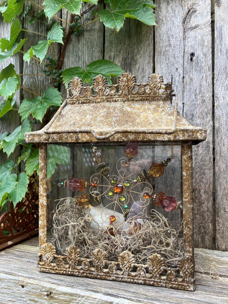 Puede incluir: Una linterna decorativa rectangular de vidrio y metal en color dorado envejecido. La linterna presenta detalles ornamentados y contiene un arreglo floral de alambre y cuentas, y musgo seco. El fondo muestra tablones de madera y vegetaci&oacute;n trepadora.