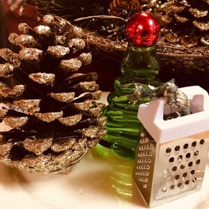 May include: A gold pine cone with glitter, a green glass bottle with a red top, and a small silver grater with a white handle.